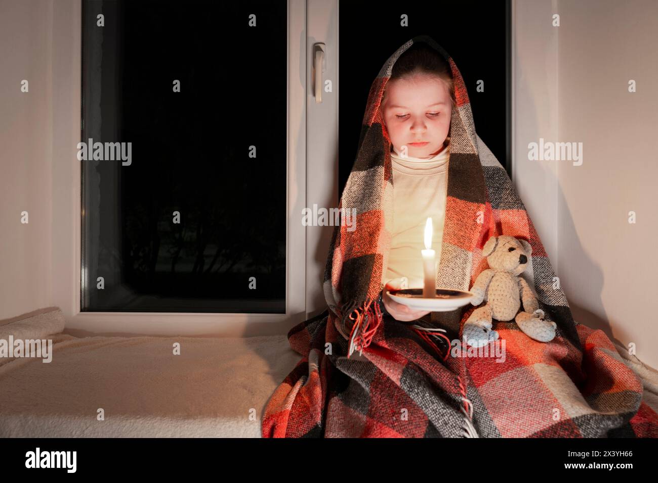 Blackout. A girl, wrapped in a blanket, sits on a windowsill near the window in a dark room and holds a teddy bear and a burning candle in her hands Stock Photo