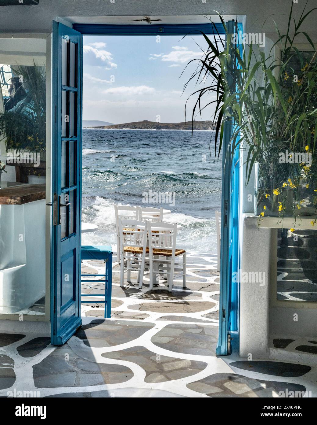 Blue doors wide open to the ocean air and sunlight, with the surf washing onto a patio with chairs; Mykonos, Greece Stock Photo