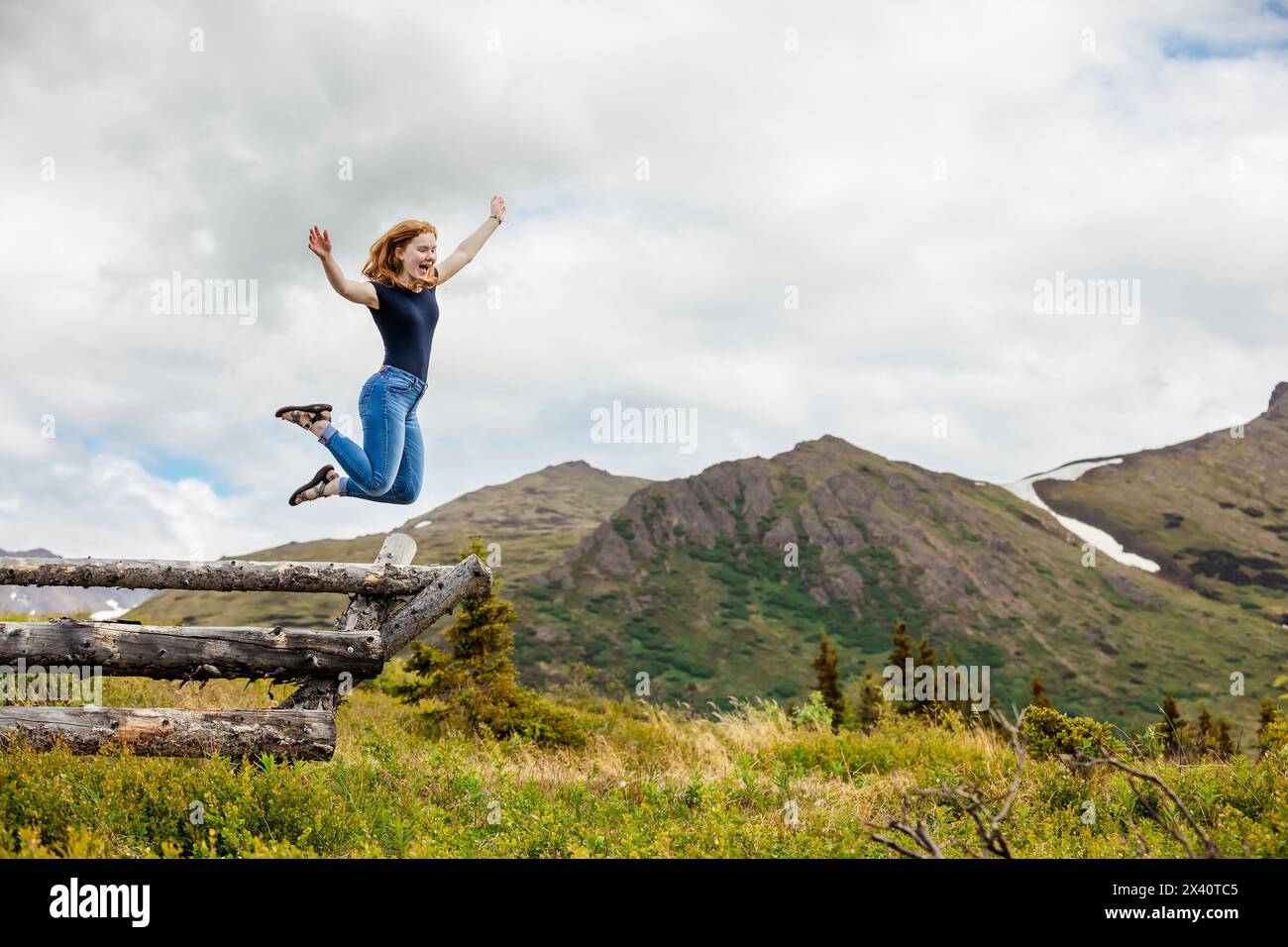 Girl jumps from a wooden rail fence with a gleeful expression, with view of the mountains in the background Stock Photo