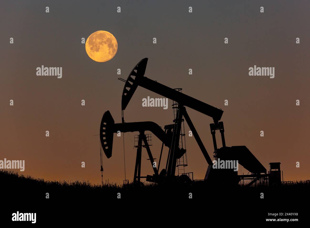 Silhouette of two pumpjacks with a glowing warm sky and full moon, West of Airdrie; Alberta, Canada Stock Photo