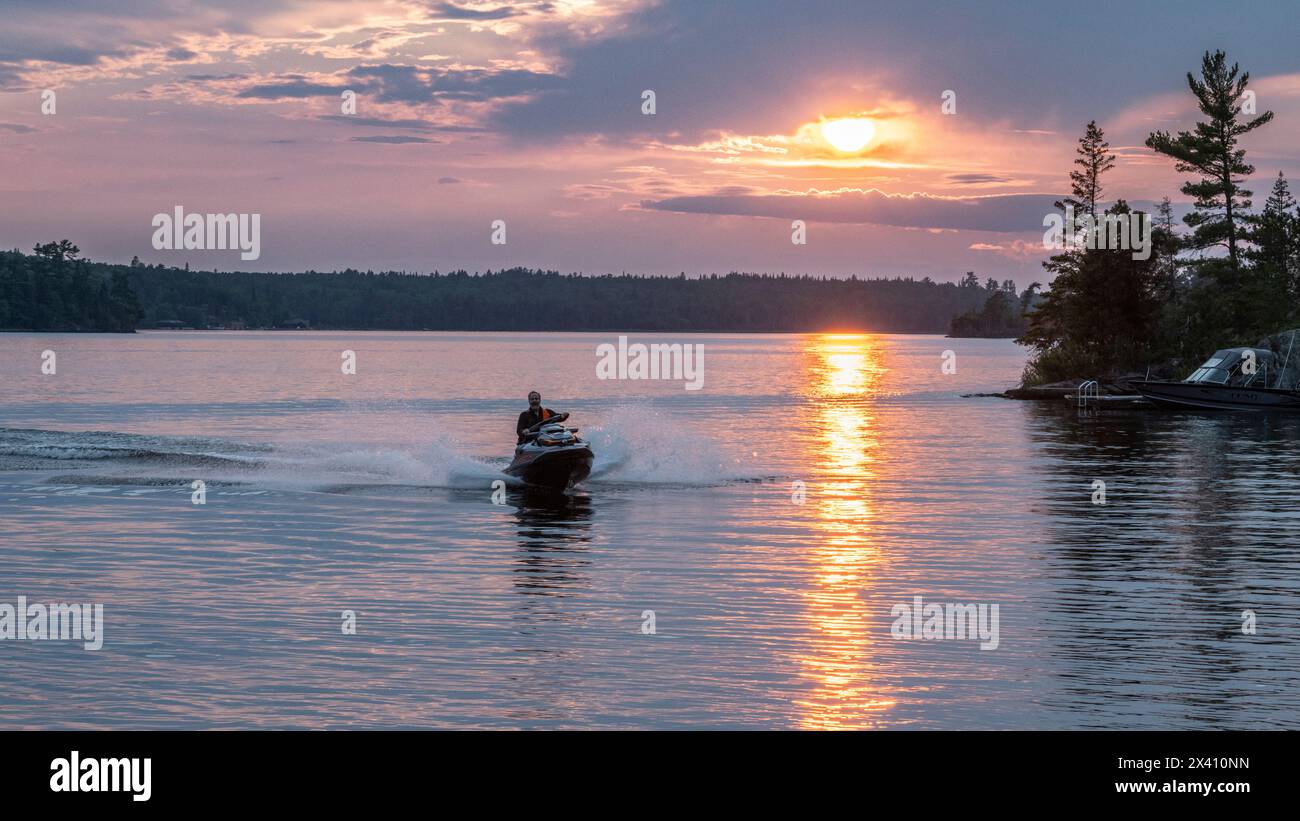 Man rides a motorized personal watercraft on a lake at sunset, with a golden sunbeam reflected on the tranquil water Stock Photo