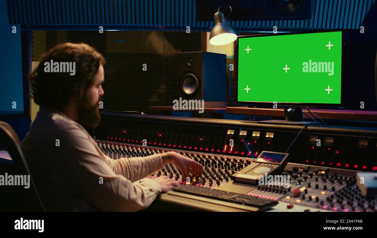 Audio expert editing tracks by following online lesson on greenscreen pc, mixing new tunes together to create a song. Sound engineer working in control room to record and process music. Camera B. Stock Photo
