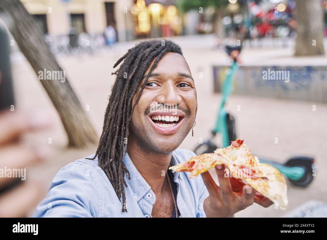 attractive man with braids holding a slice of pizza in his hand while taking a photo with his smart phone on the street Stock Photo