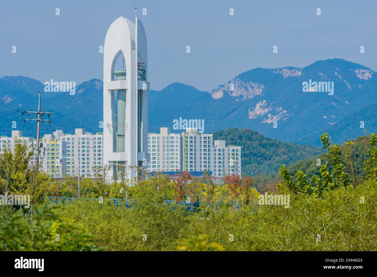Tall white tower with three sides and cylindrical metal column in middle standing in woodland area with high rise apartments and mountains in Stock Photo