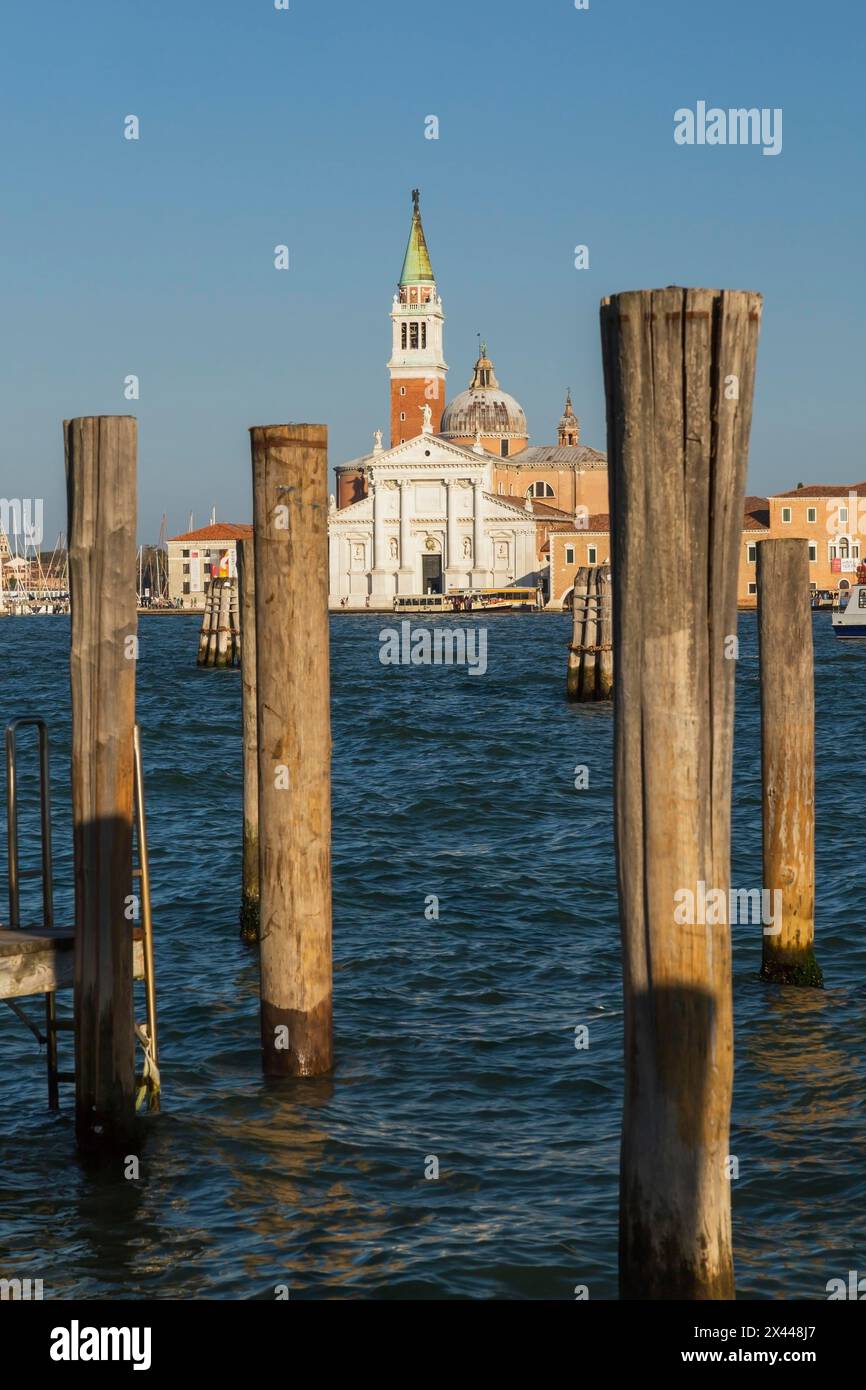 Wooden mooring posts canal lane markers in Venetian lagoon and 16th century Benedictine church of San Giorgio Maggiore with bell tower on San Giorgio Stock Photo
