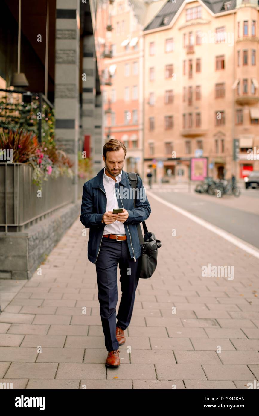 Full length of businessman using smart phone while walking on sidewalk in city Stock Photo