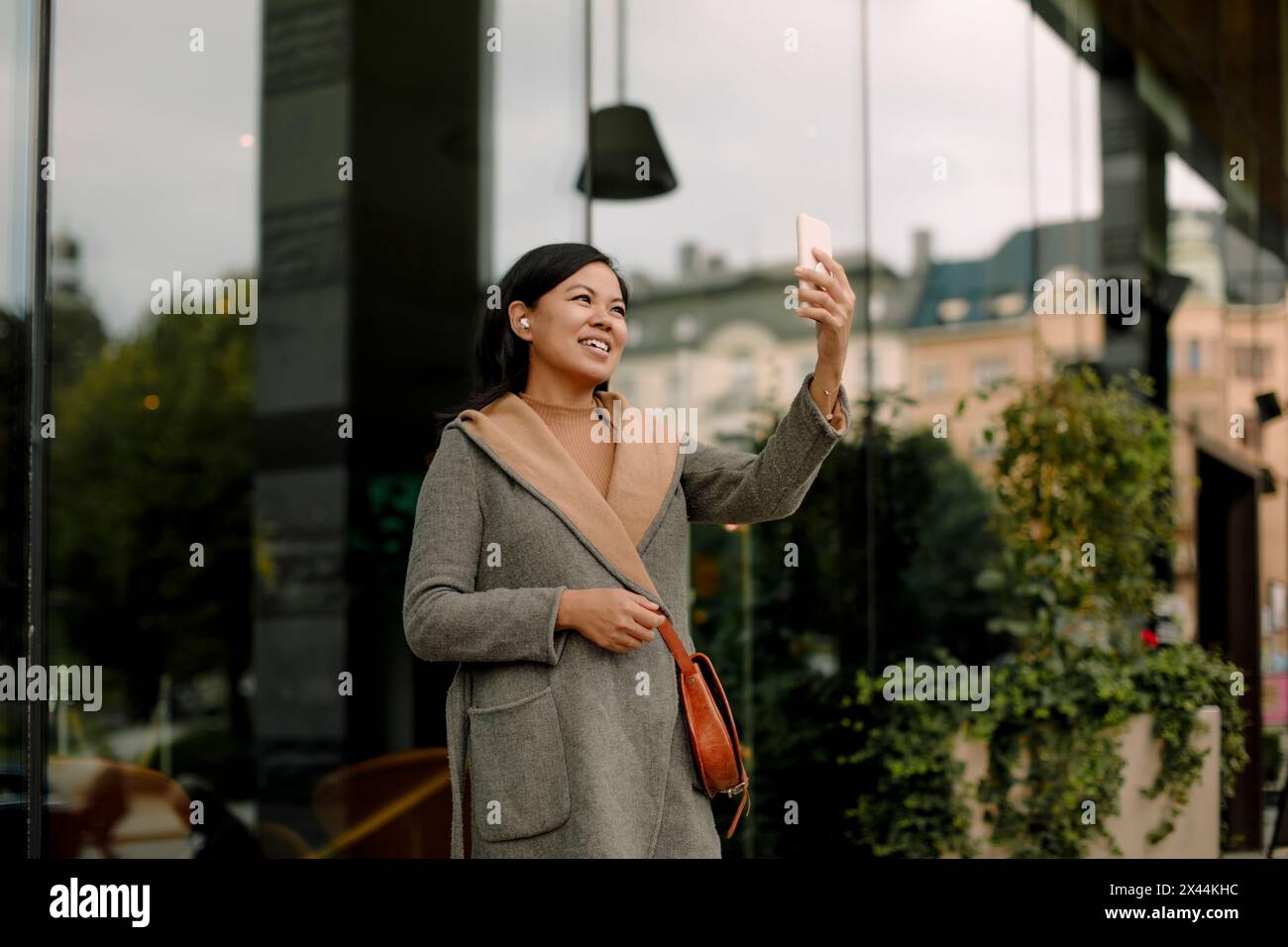 Smiling businesswoman on video call using smart phone while standing against glass building Stock Photo