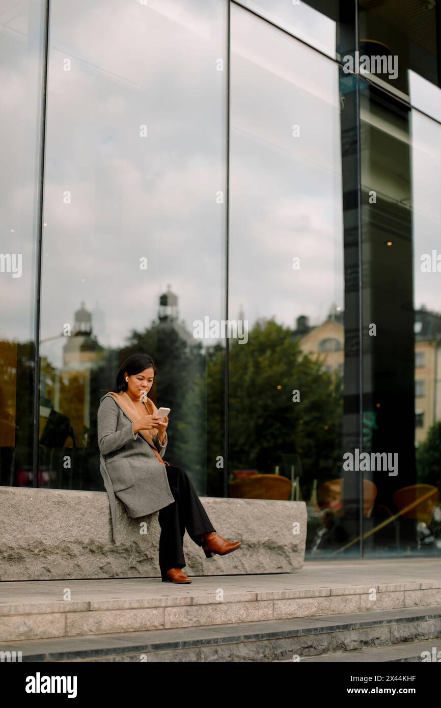 Full length of businesswoman using smart phone while sitting on bench Stock Photo