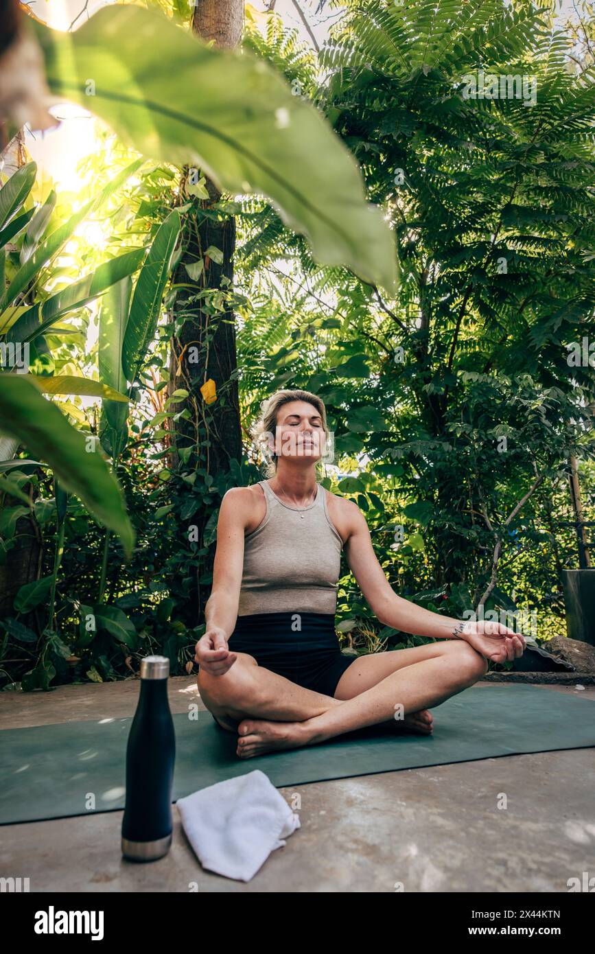 Woman practicing Lotus position while sitting on yoga mat near plants ...