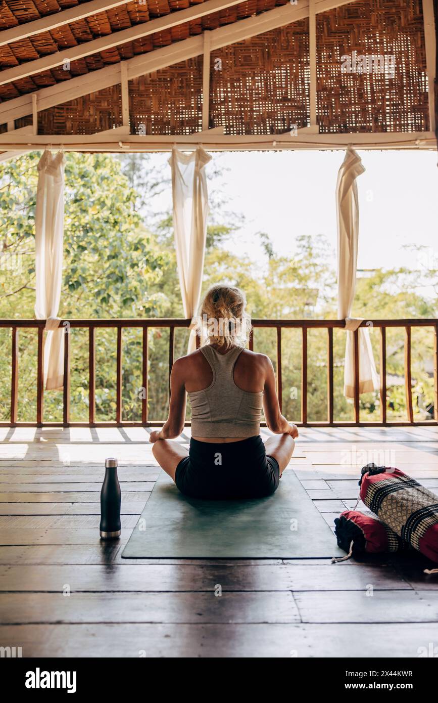 Rear view of woman doing yoga while sitting on exercise mat over wooden floor at wellness resort Stock Photo