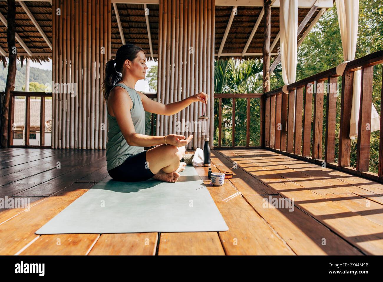 Side view of woman holding Tibetan bells and meditating while sitting on yoga mat at wellness resort Stock Photo