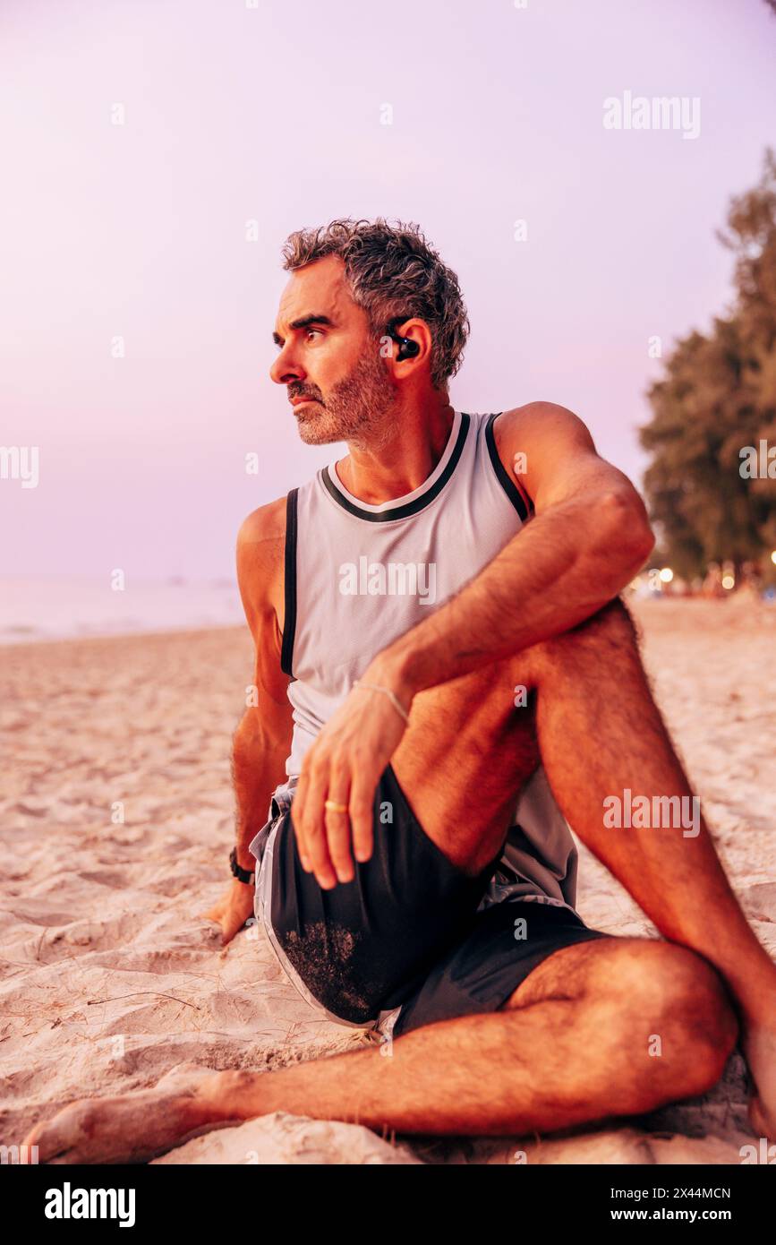 Mature man doing stretching while sitting on sand at beach Stock Photo