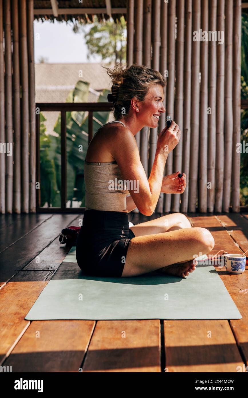 Side view of smiling woman smelling oil while sitting on yoga mat at wellness resort Stock Photo