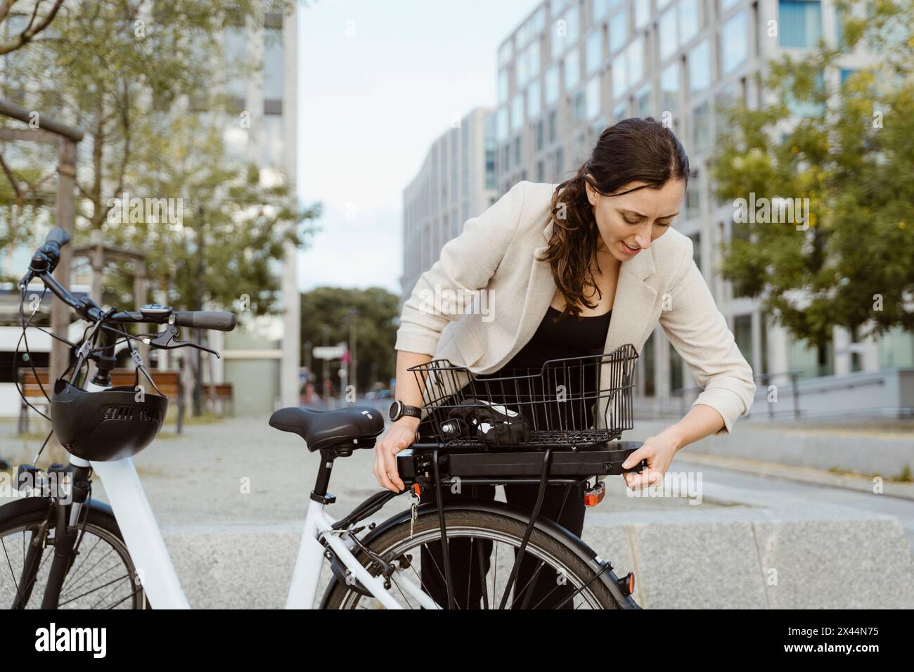 Businesswoman removing battery from electric cycle while standing against office building in city Stock Photo