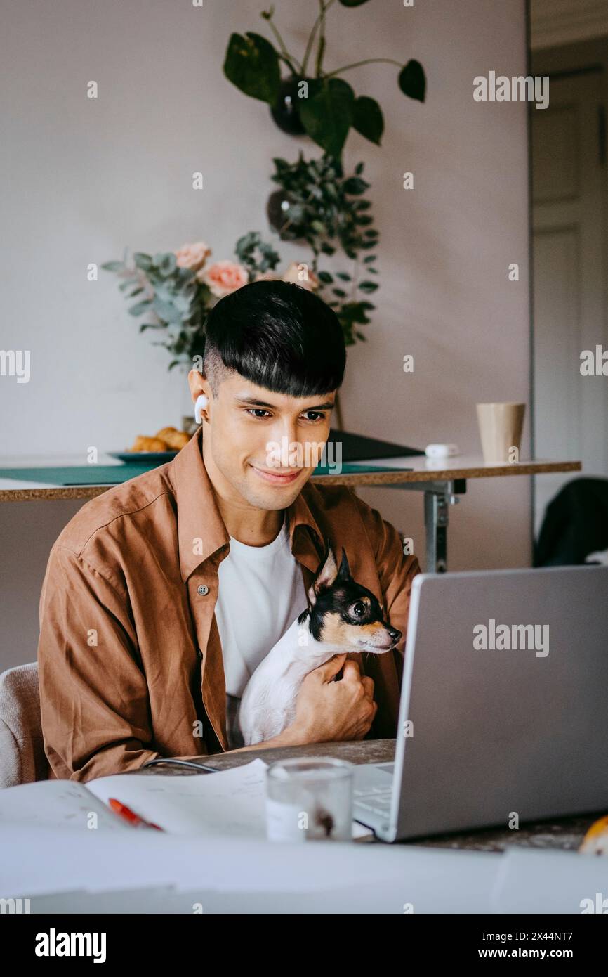 Smiling freelancer sitting with dog and working on laptop at home office Stock Photo