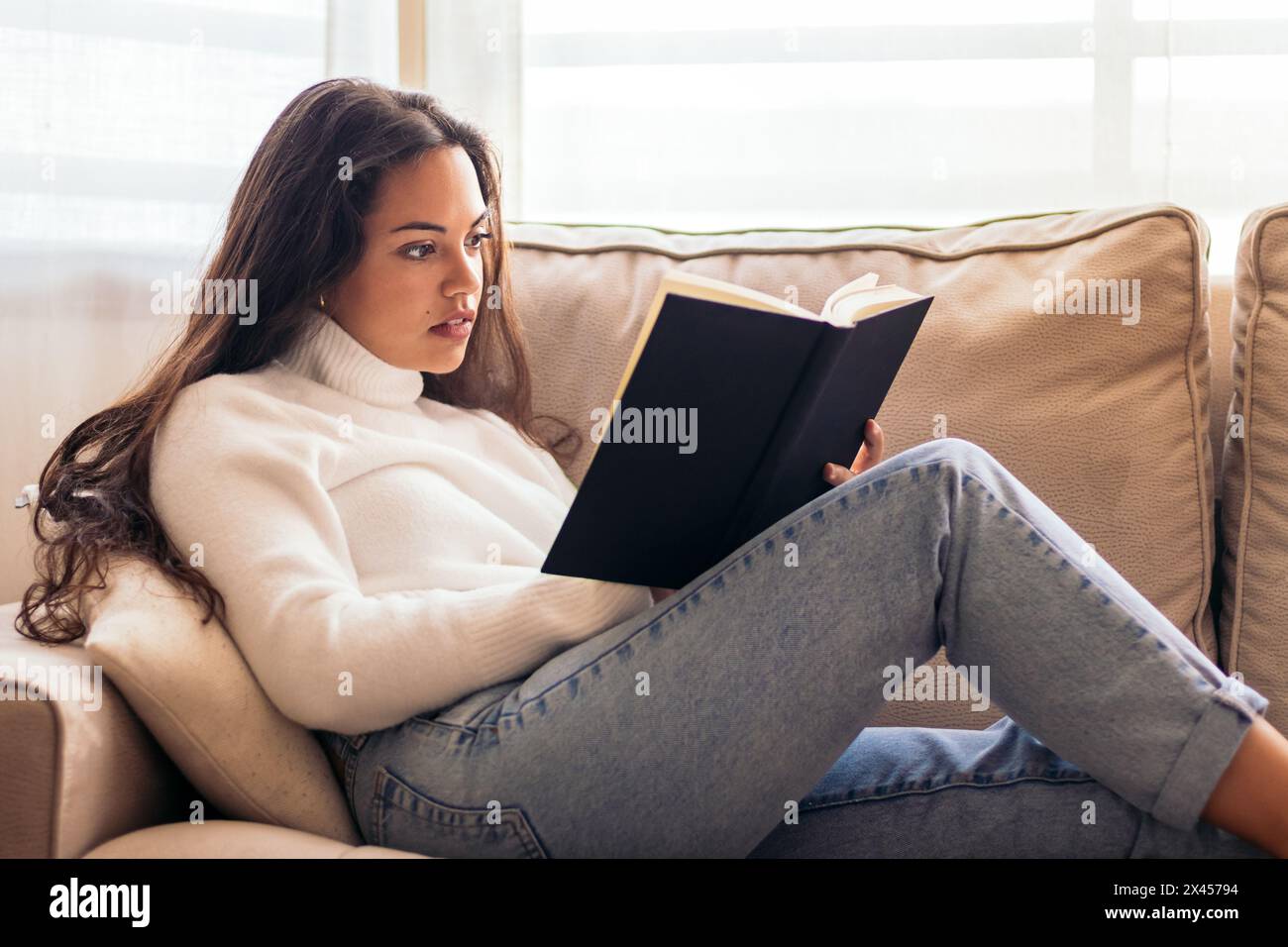 Young woman sitting on her couch at home, engrossed in a book. reading, relaxation, inner peace, and a cozy home Stock Photo