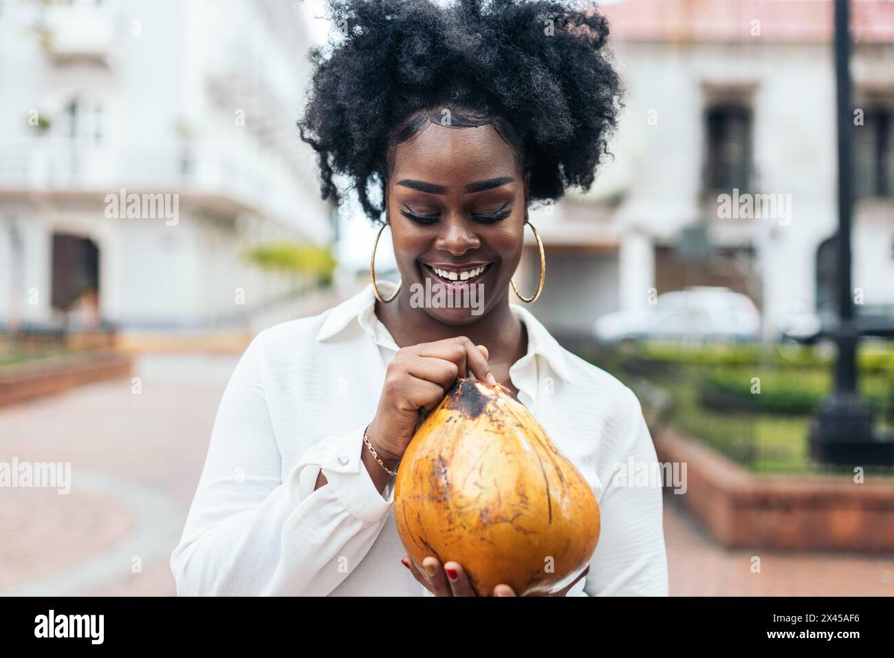 Portrait of an afro-haired black Latin American woman with large earrings holding and drinking a coconut. She is happy and smiling. Stock Photo