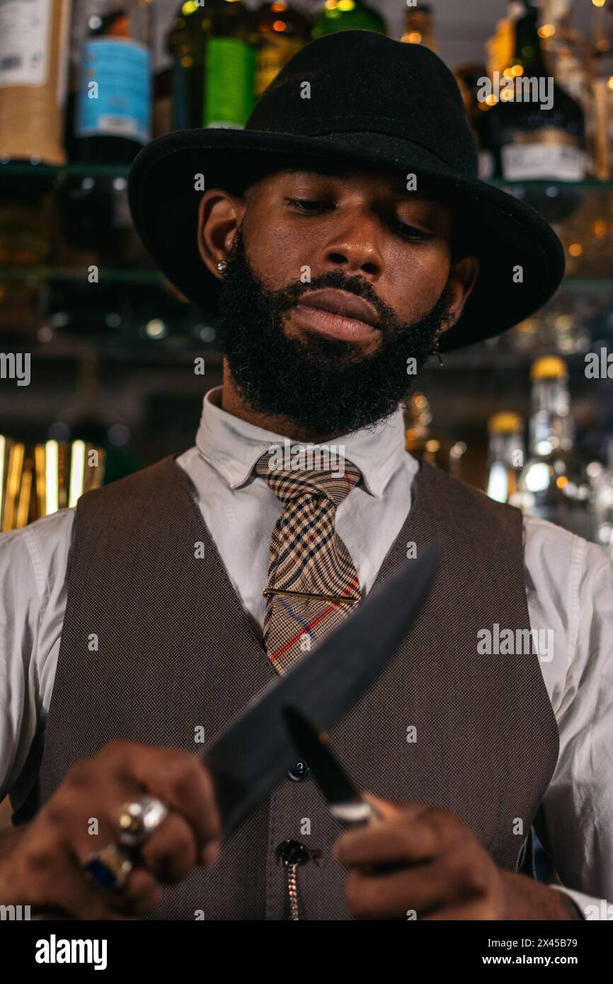 Close-up of a stylish black bartender in a hat sharpening his knives Stock Photo
