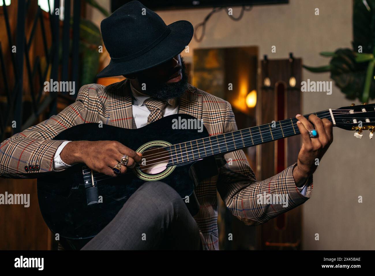 Stylish black man in hat and classic outfit playing guitar indoors Stock Photo