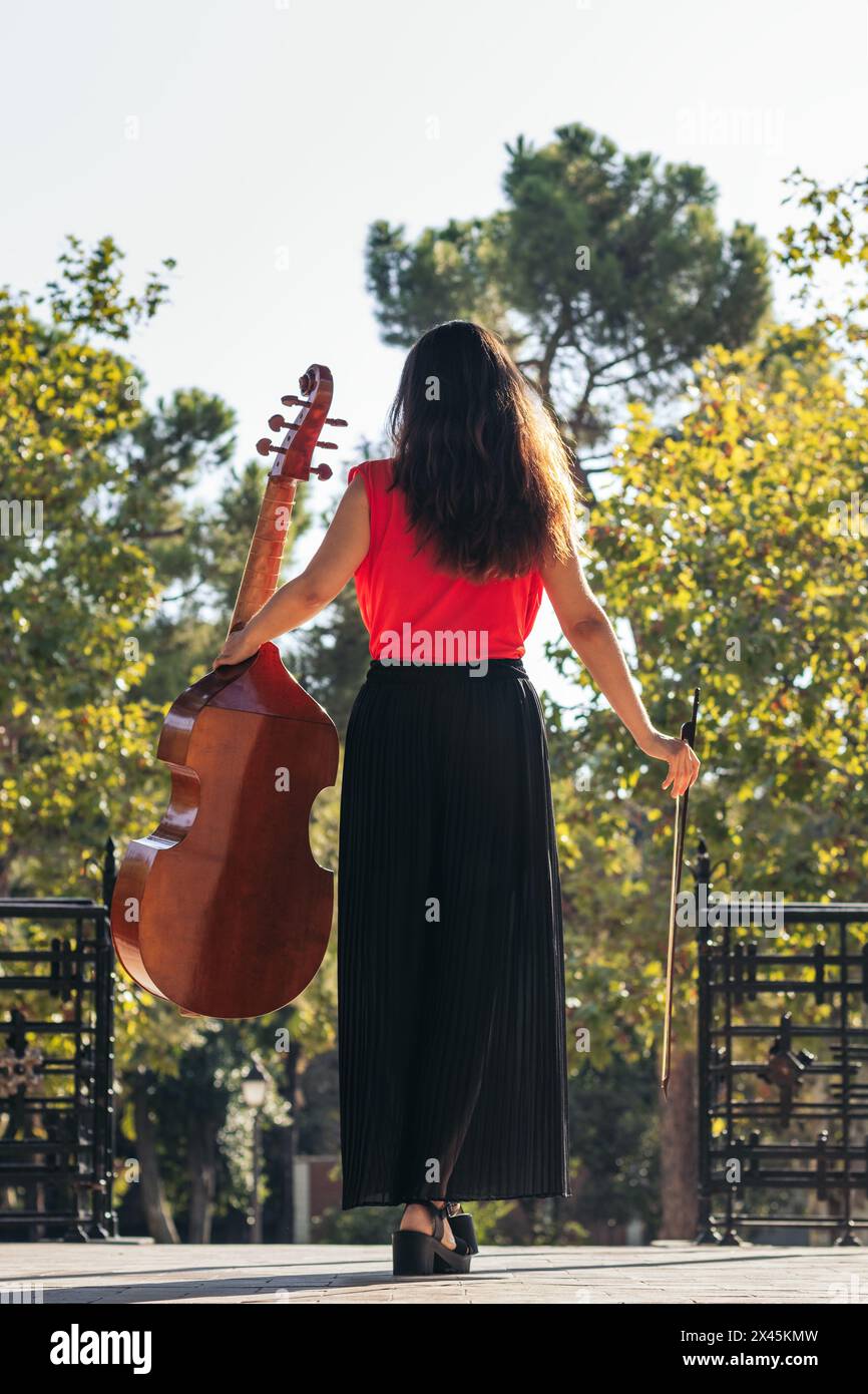 A rear view of a female musician standing, holding her cello, on a stage in the gardens of an ancient European palace. Stock Photo