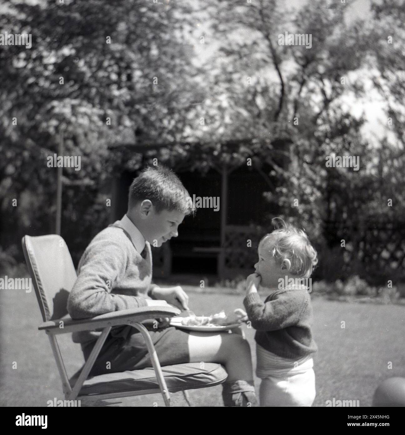 1960s, historical, summertime and two young boys, brothers, the older one sitting outside on a lawn having a meal on his lap after school, his infant brother standing opposite him.....interested in his food! Stock Photo