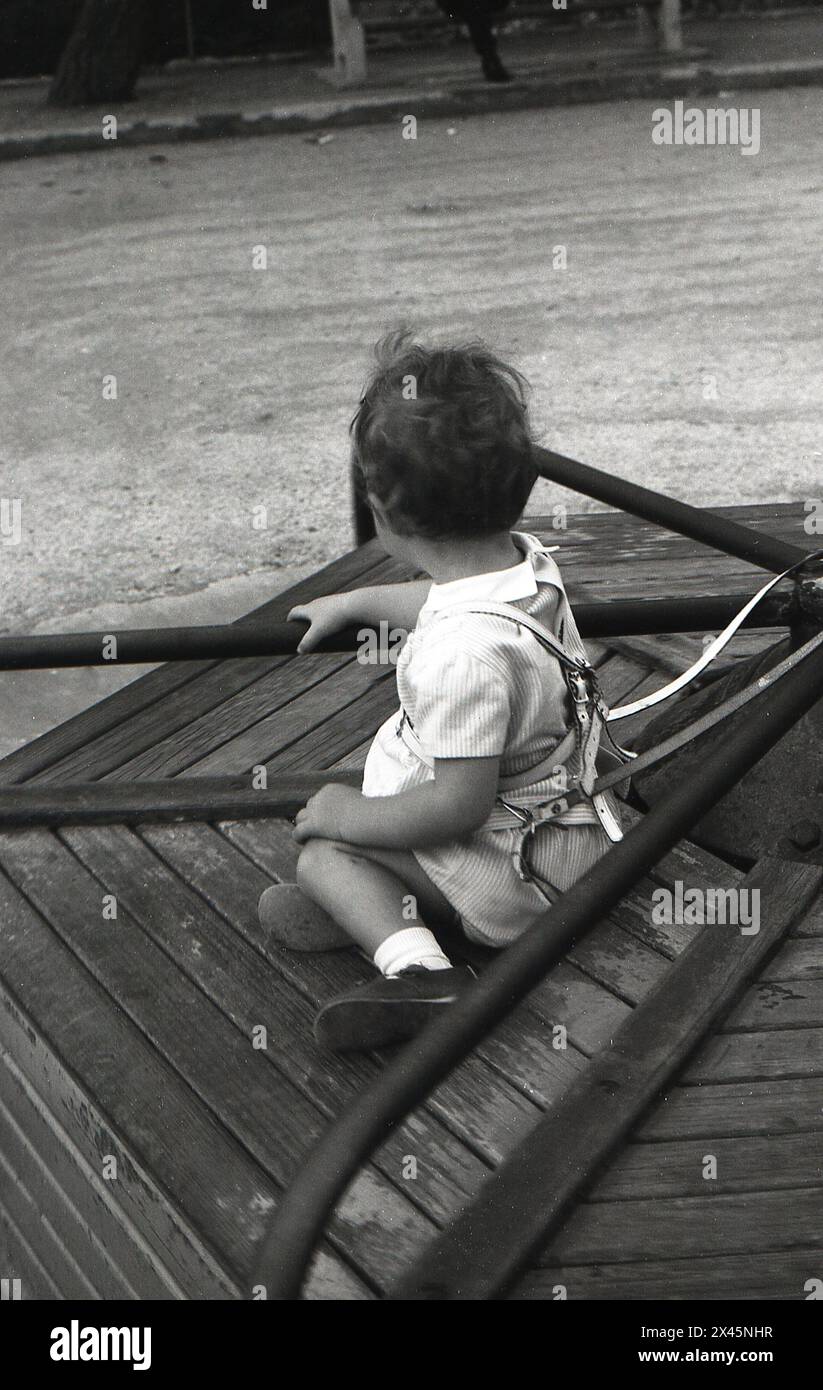 1960s, historical, an infant child, wearing a leather safety harness, sitting having a ride on the raised wooden platform of a traditional metal playground roundabout. Stock Photo