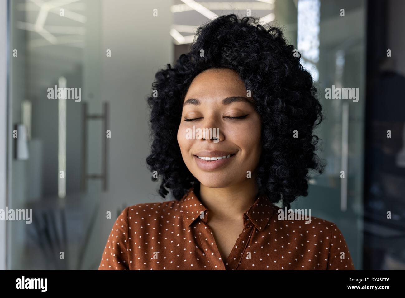 A young African-American woman with curly hair, dressed in a polka dot shirt, closes her eyes and smiles, enjoying a serene moment in a sunlit modern office environment. Stock Photo