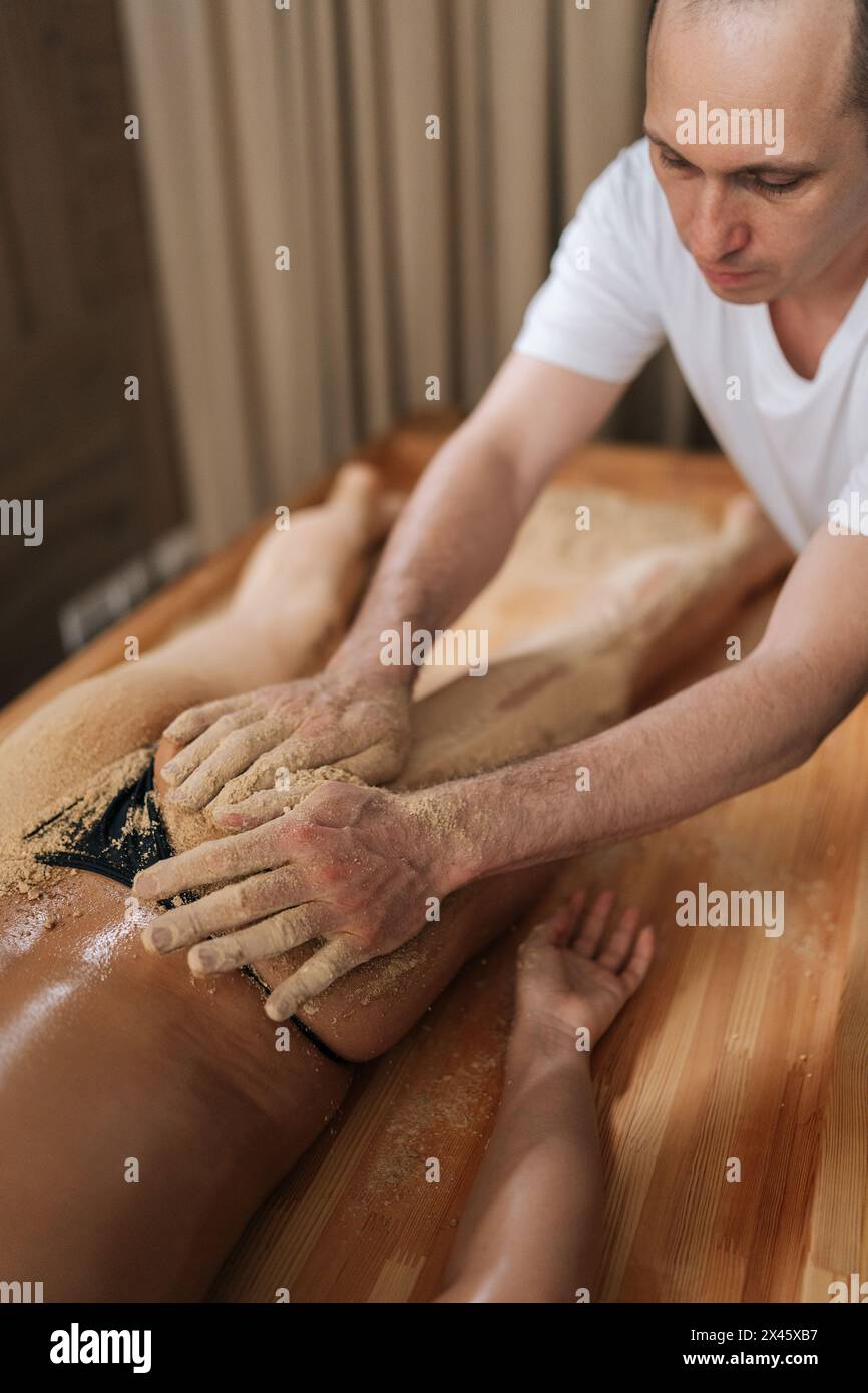 Vertical shot of male massage therapist scrubbing massaging tanned buttocks  with warm sand of female client lying on table in wellness center Stock  Photo - Alamy