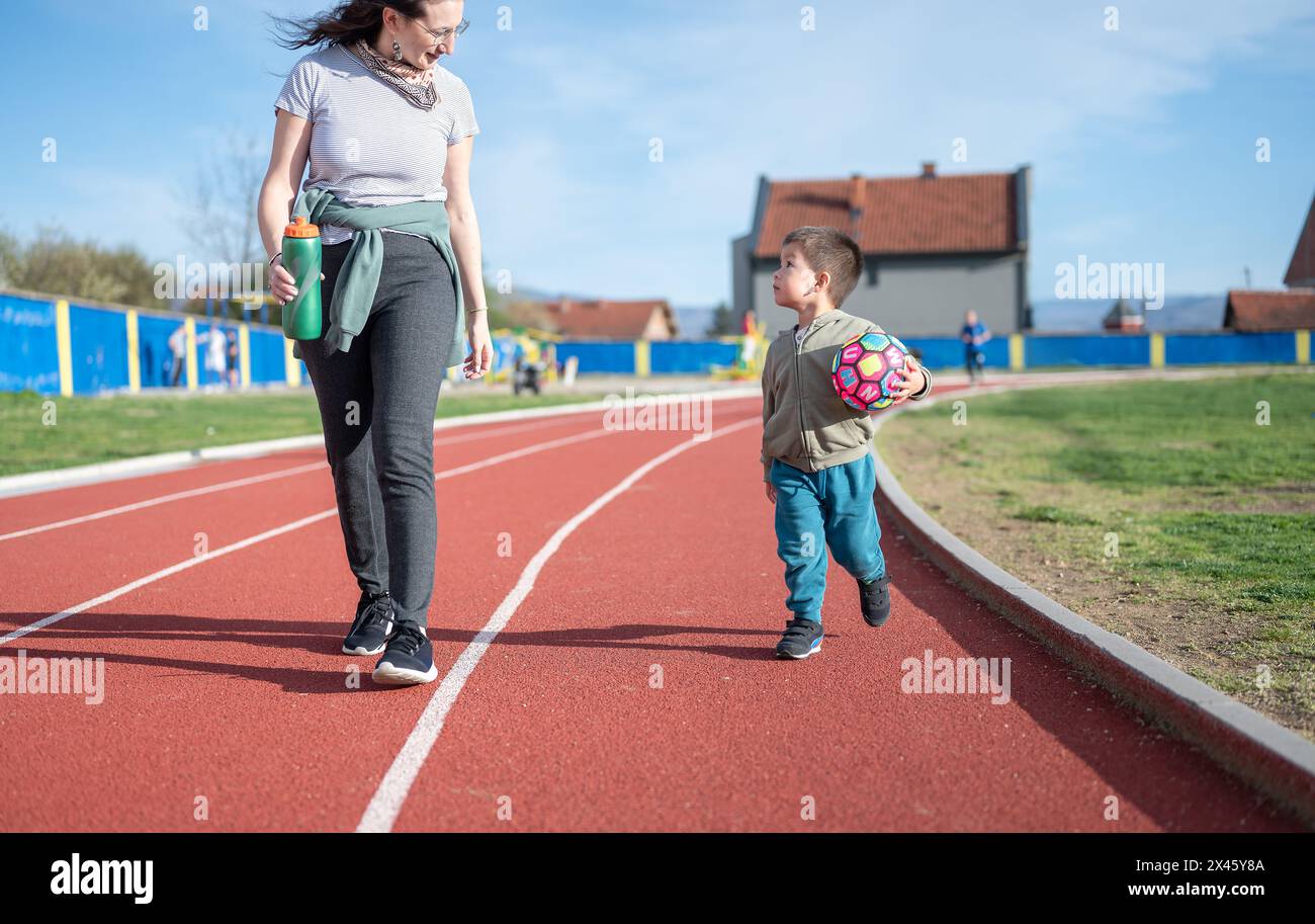 A 3 year old toddler excitedly runs with a soccer ball on a track field with his aunt, indicating an early love for football sports The image captures Stock Photo