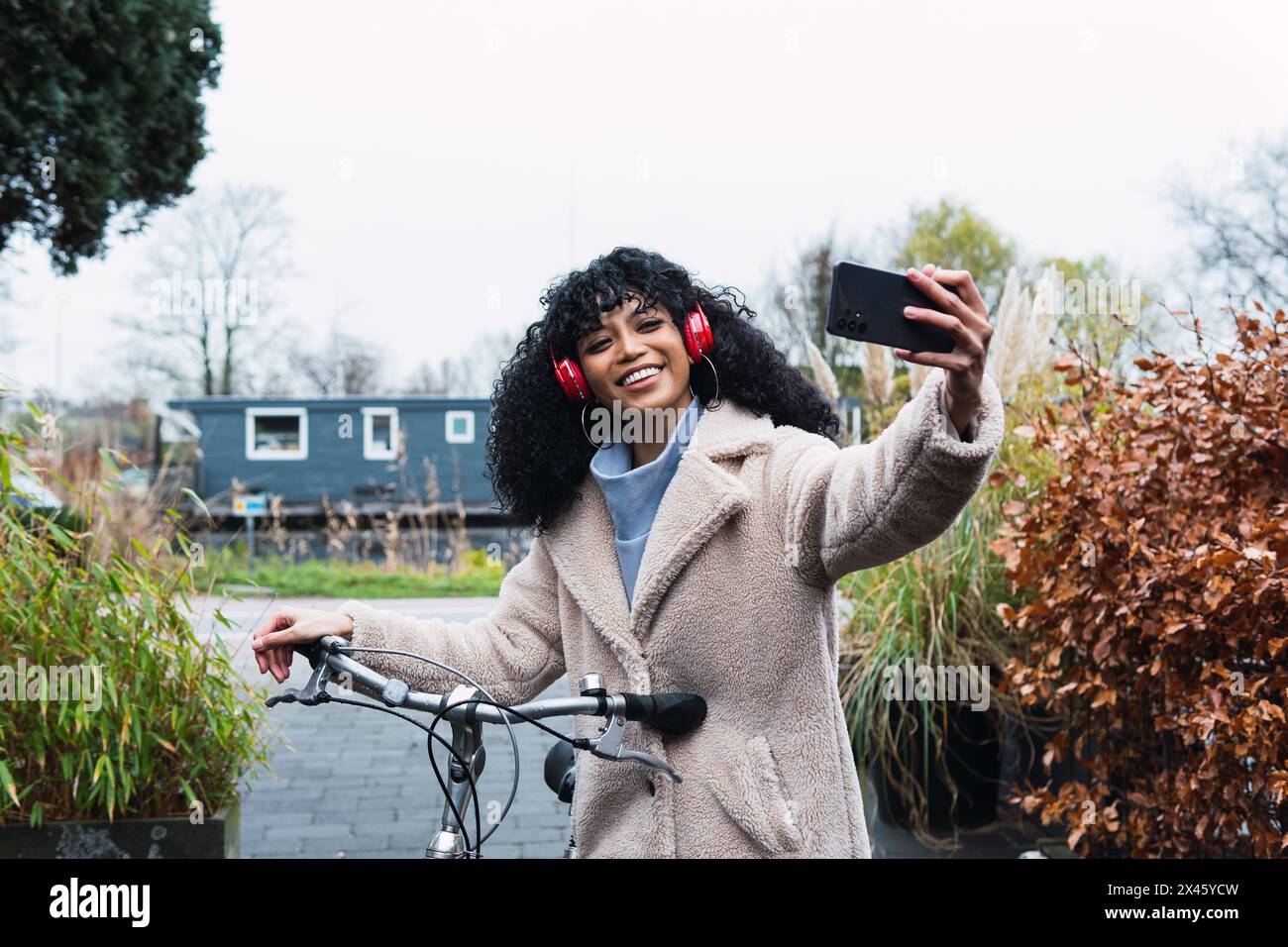 Cheerful young African American female with curly dark hair in warm coat walking with bicycle while taking selfie and listening to music in headphones Stock Photo