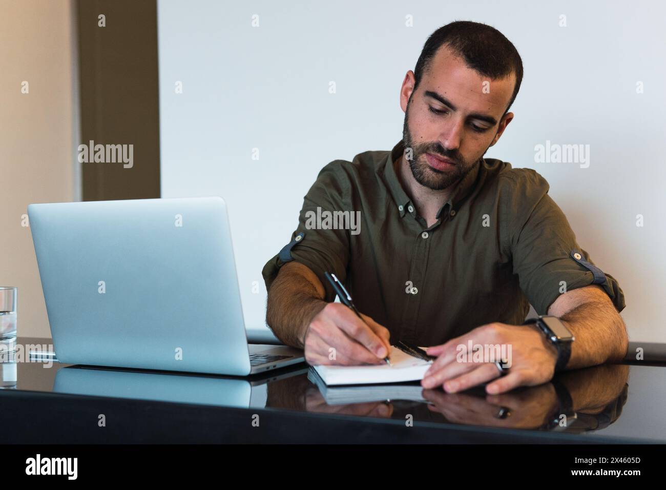 Focused male taking notes in notepad while sitting at table with modern netbook in light room during work time Stock Photo