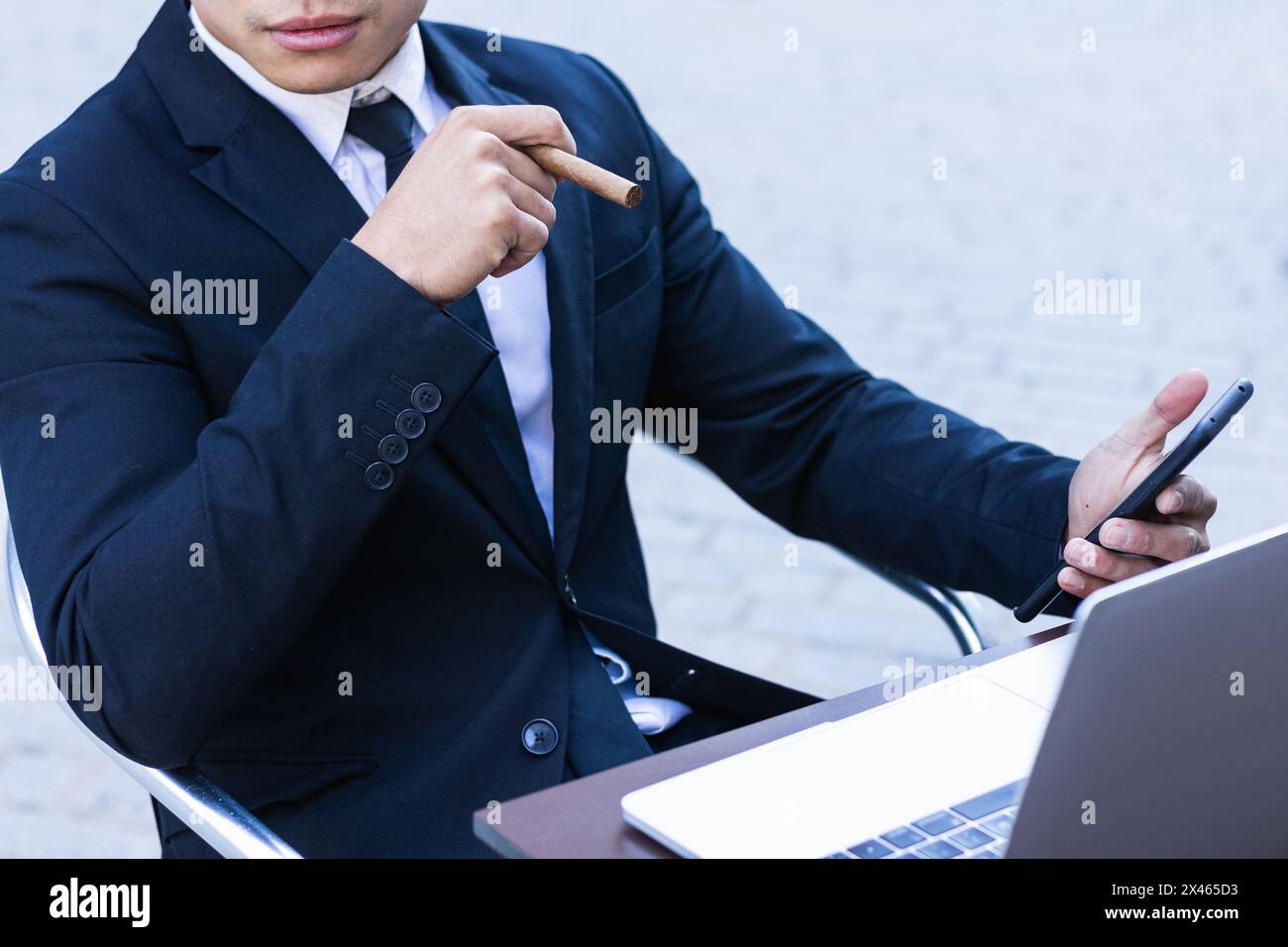Confident Asian male entrepreneur sitting at table in cafe and smoking cigar while browsing smartphone Stock Photo