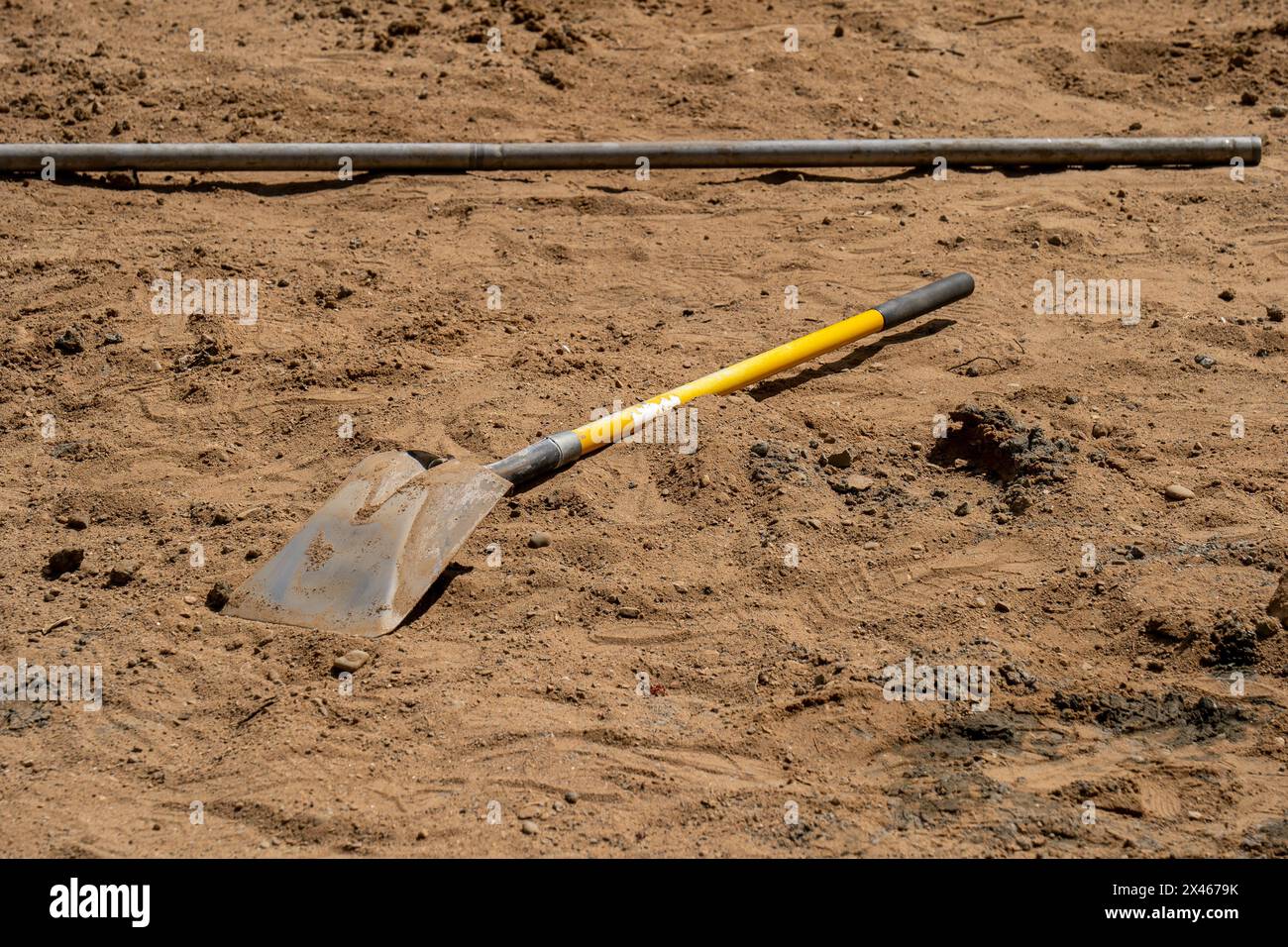 A shovel with a yellow handle laying in the brown dirt, near a metal pipe, at the construction site. A worker's tool for cleaning up and moving soil. Stock Photo