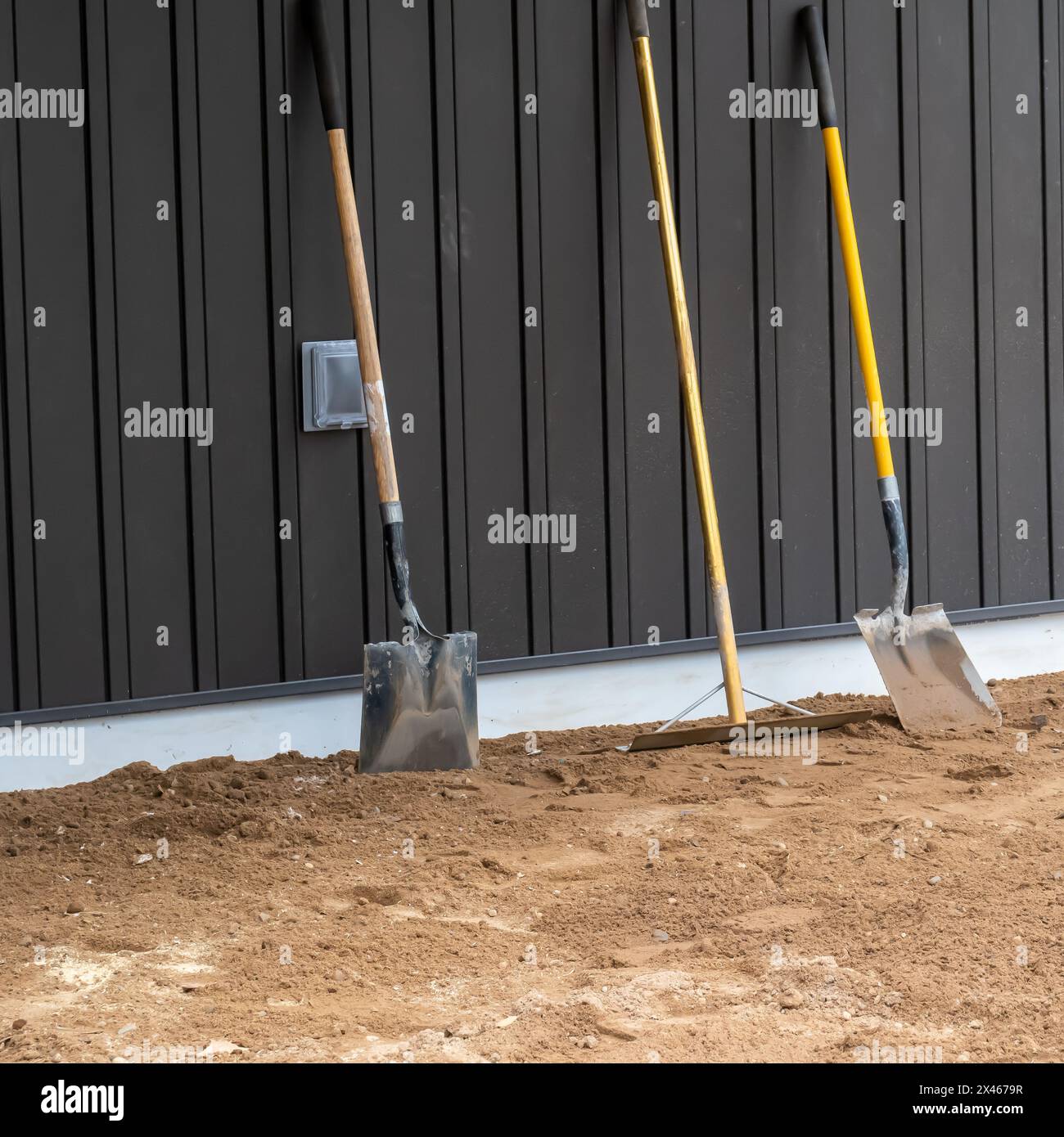 Dirty, used shovels and a rake stand upright on the dirt and lean against the board and batton siding of a wall at a construction site. Stock Photo