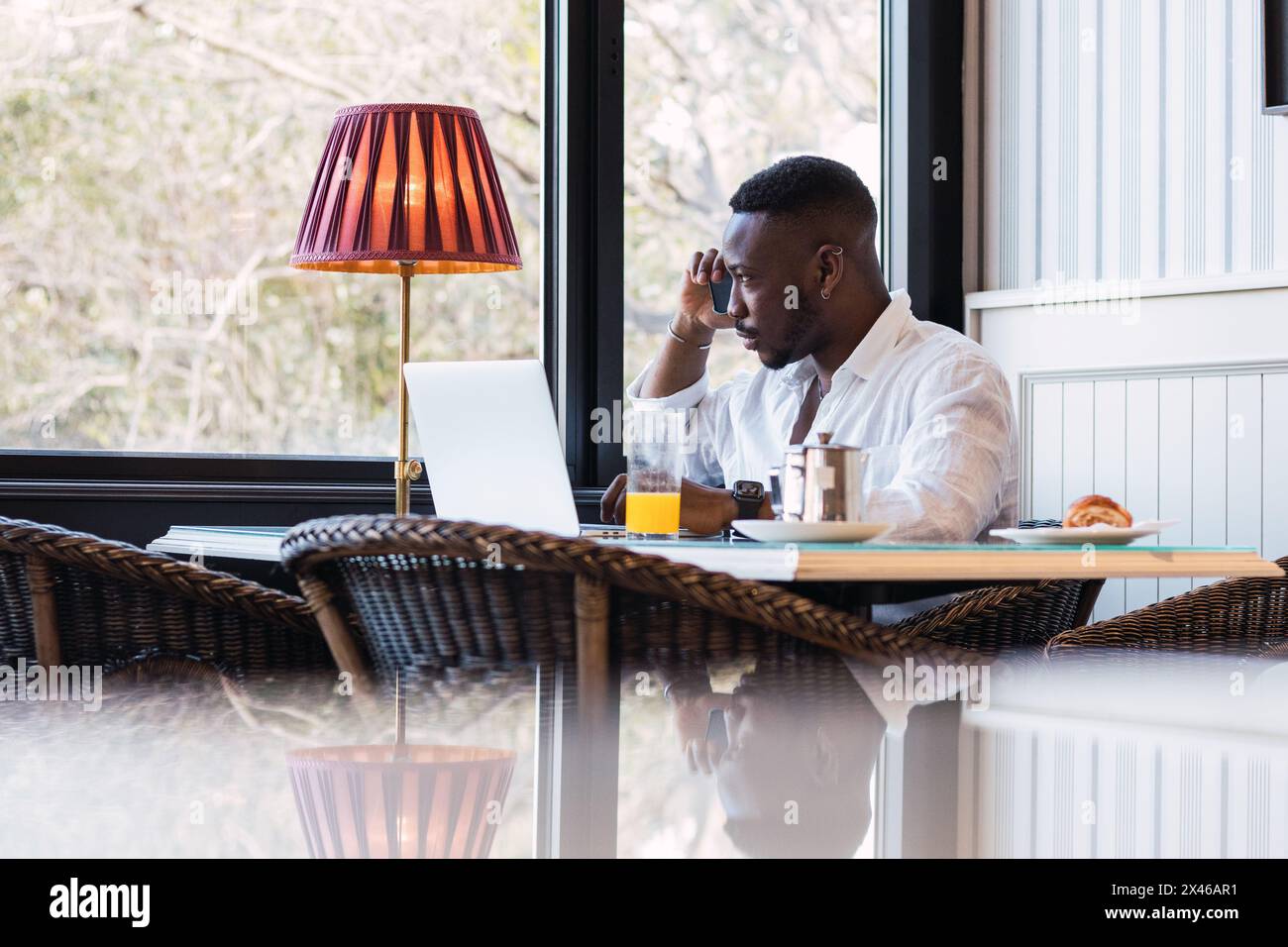 Serious adult African American businessman sitting at table with laptop and discussing business issue during phone call while having breakfast in cozy Stock Photo