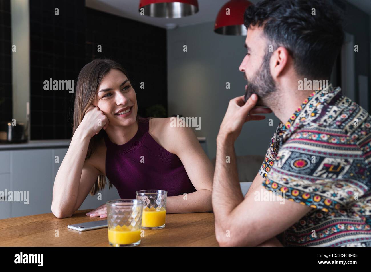Positive young couple in casual clothes having conversation and enjoying fresh orange juice while sitting at dining table in kitchen Stock Photo