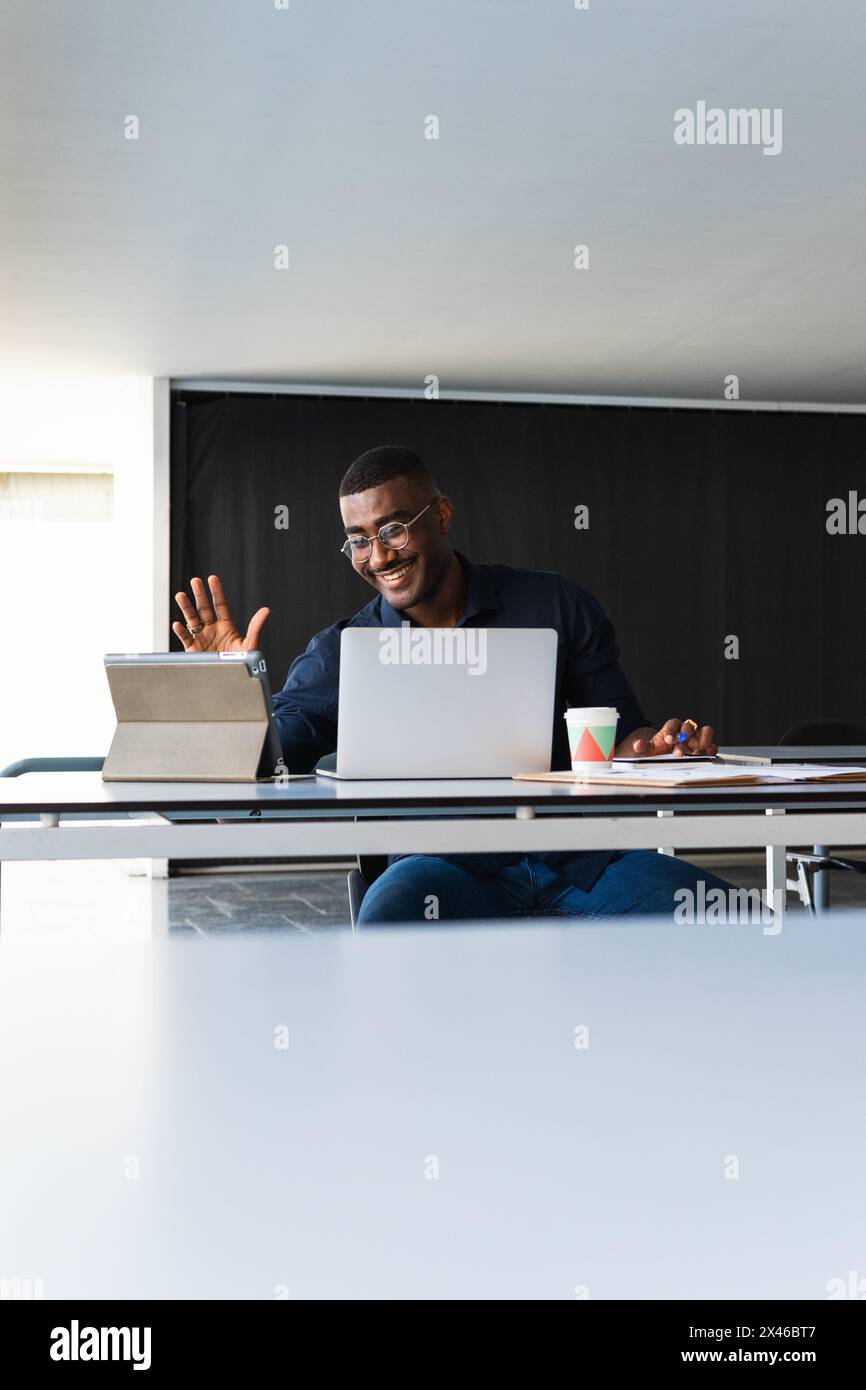 Positive African American adult male in formal clothes and eyeglasses sitting at table while having video call on tablet near netbook and documents wi Stock Photo
