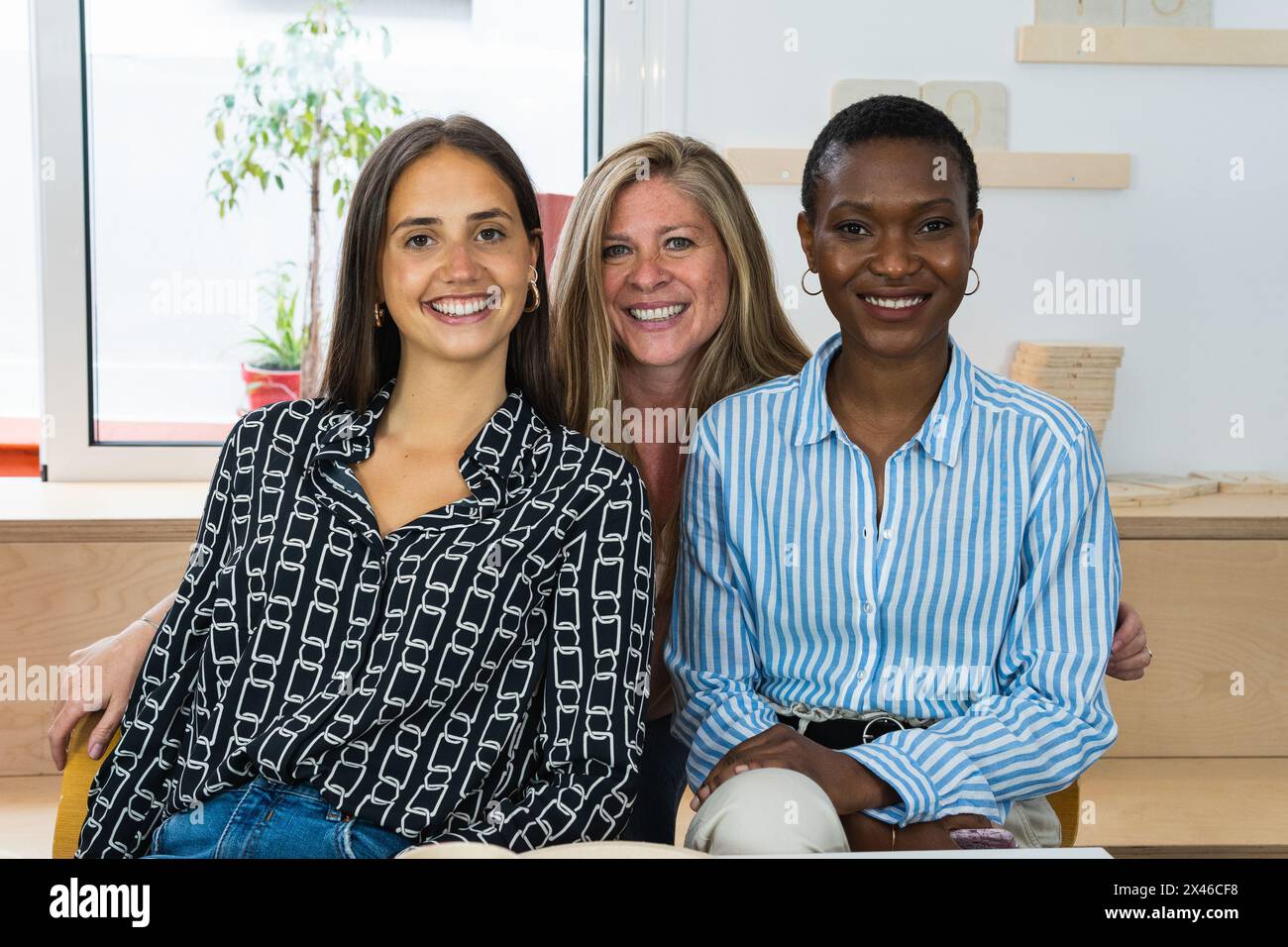 Delighted multiracial female colleagues looking at camera and smiling while sitting in workplace together Stock Photo