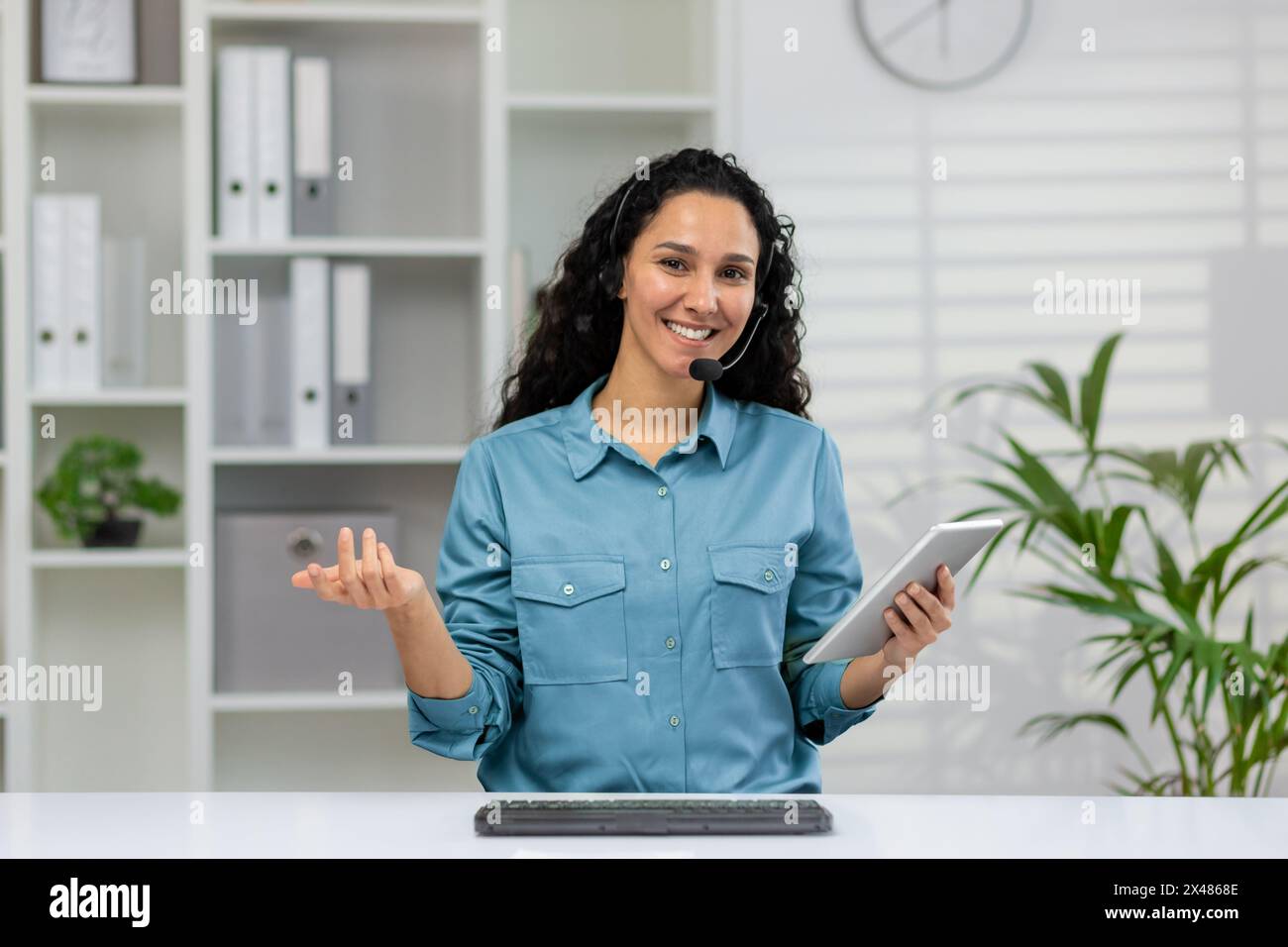 A smiling woman in a blue shirt wearing a headset works as a customer service representative. She gestures openly with a tablet in her hand, in a brig Stock Photo