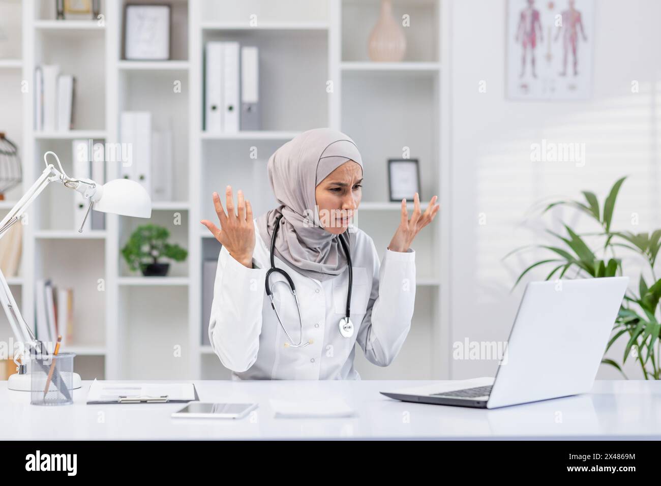 Professional female doctor wearing a hijab looks frustrated while working on her laptop in a modern clinic office. Perfect image for healthcare challe Stock Photo