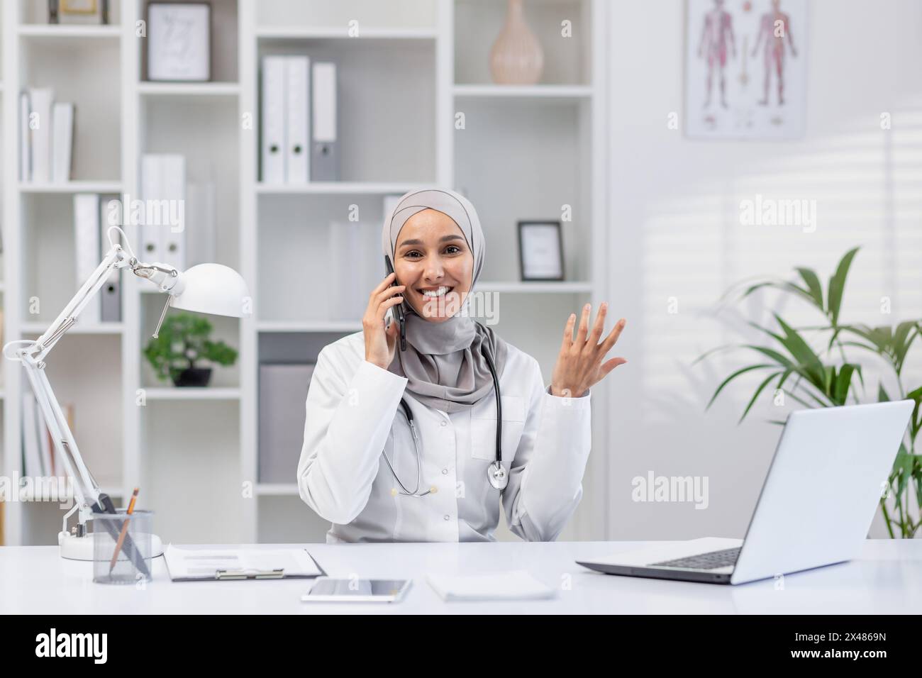 Smiling Muslim female doctor in a hijab having a conversation on the phone while sitting at her desk in a well-lit clinic office, looking satisfied. Stock Photo