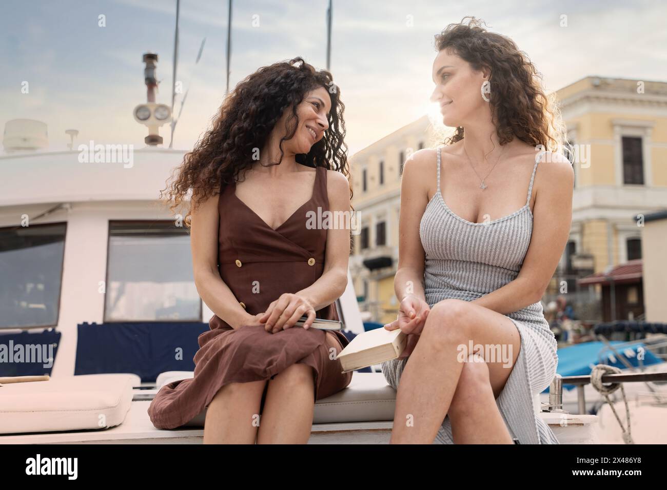 Two women - conversing on yacht - urban backdrop, casual outfits, relaxing day - friendship, lifestyle, leisure. Stock Photo