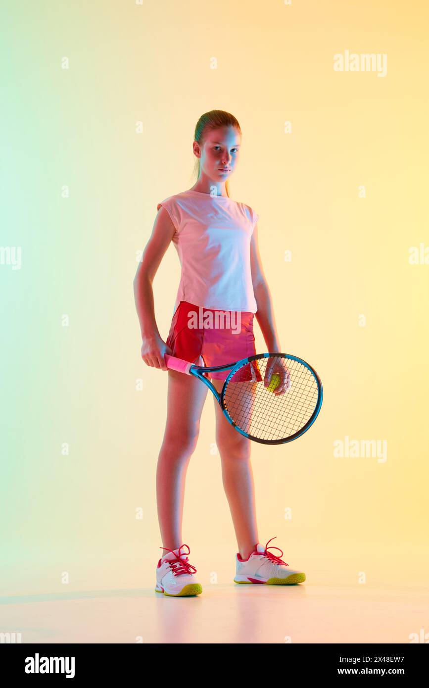Tennis player. Purposeful girl teenager and athlete with racket in pink-red sportswear posing in neon light against blue-yellow background. Stock Photo