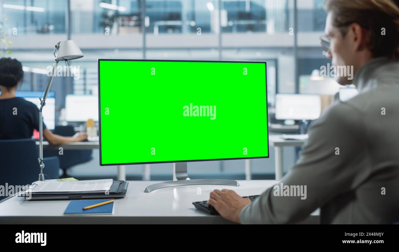 Close Up Over the Shoulder Shot of a Businessman Working on Desktop Computer with Chroma Key Green Screen Mock Up Display. Digital Projects Manager Typing Data, Using Keyboard and Mouse. Stock Photo