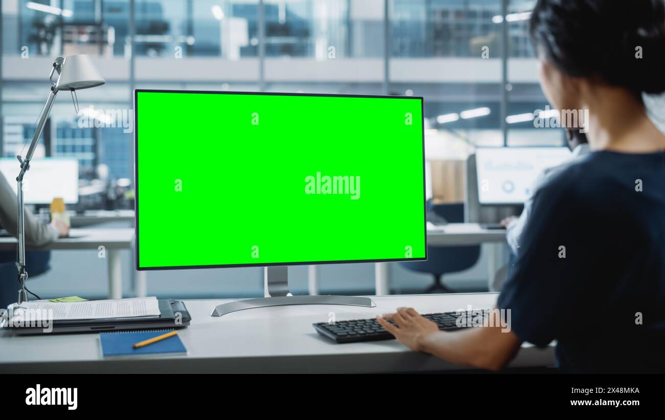 Close Up Over the Shoulder Shot of a Businesswoman Working on Desktop Computer with Chroma Key Green Screen Mock Up Display. Digital Projects Manager Typing Data, Using Keyboard and Mouse. Stock Photo
