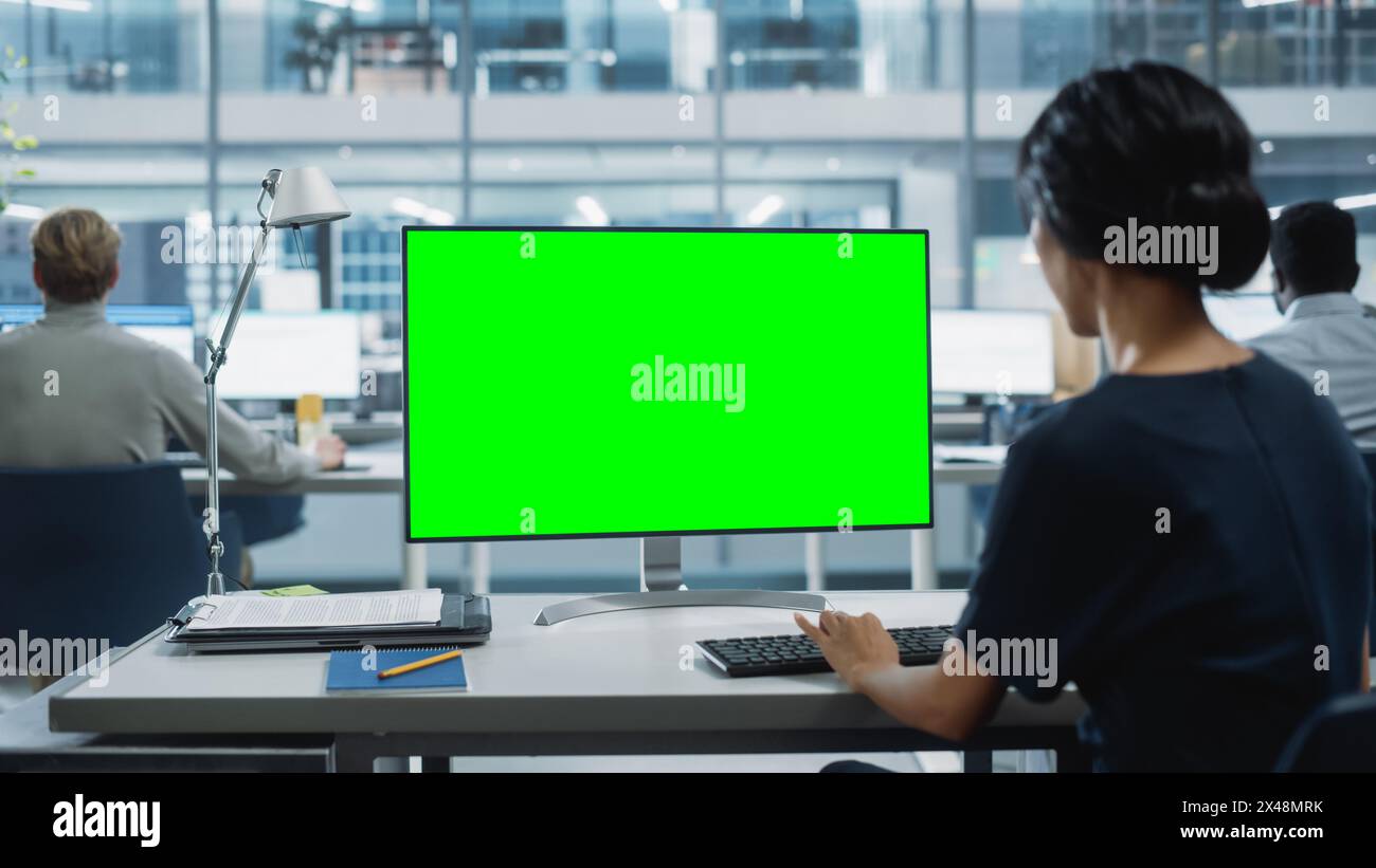 Close Up Over the Shoulder Shot of a Businesswoman Working on Desktop Computer with Chroma Key Green Screen Mock Up Display. Digital Projects Manager Typing Data, Using Keyboard and Mouse. Stock Photo