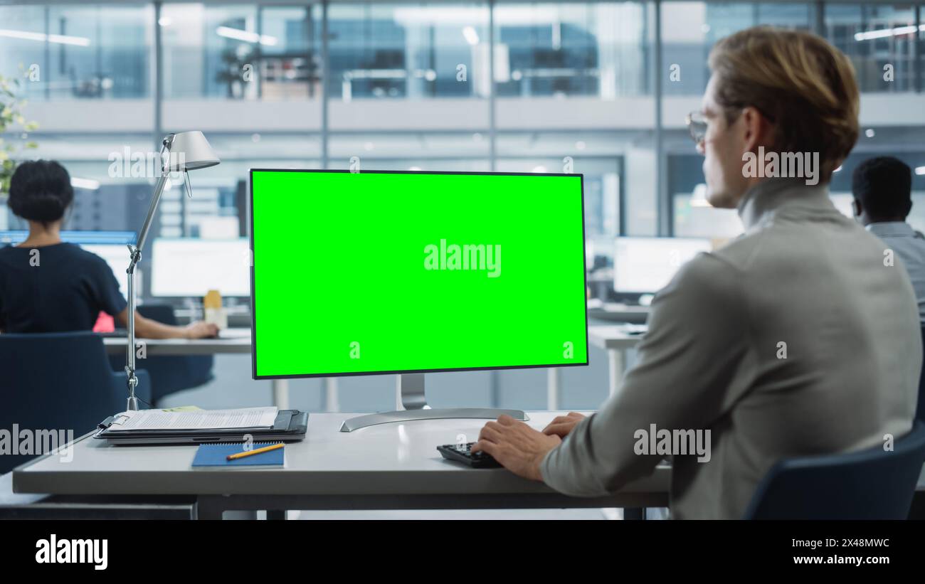 Close Up Over the Shoulder Shot of a Businessman Working on Desktop Computer with Chroma Key Green Screen Mock Up Display. Digital Projects Manager Typing Data, Using Keyboard and Mouse. Stock Photo