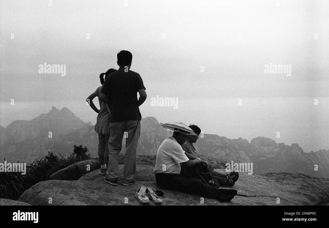 In the summer of 2014, atop Mount Lao in Qingdao, Shandong Province,A small group of individuals stands and sits atop a rocky mountain peak, overlooking a vast expanse of craggy mountains under a hazy sky. The mood is contemplative and serene as they take in the natural beauty of the landscape at the close of day. /China Stock Photo