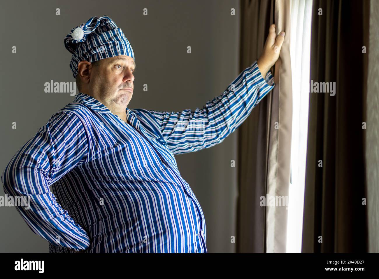 A fat man in striped pajamas looks out of a curtained window Stock Photo