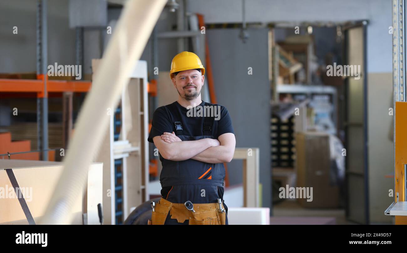 Portrait of young attractive man in work clothes and yellow helmet crossed arms smiling at shop for manufacturing furniture and details home interior Stock Photo